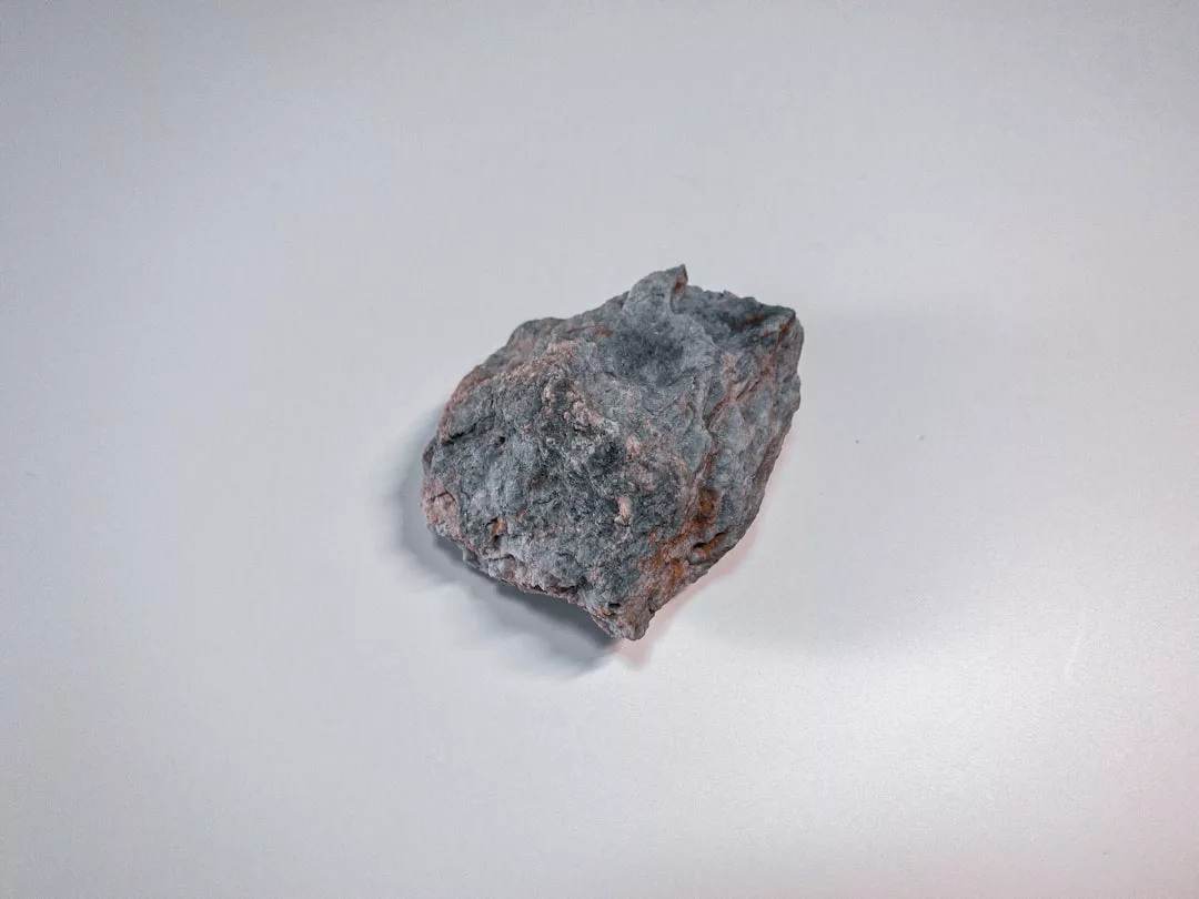 Ein einzelner rauer, grauer GLAMESiA-Stein mit ungleichmäßiger Textur und Kanten ruht auf einer glatten, hellen Oberfläche.