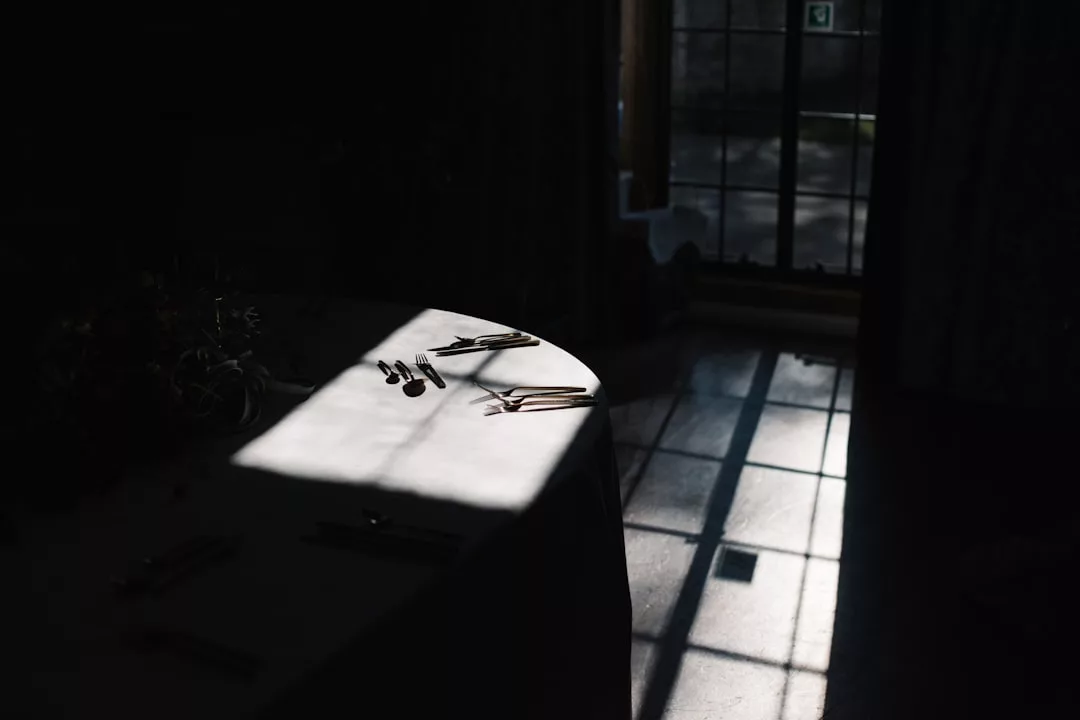 Ein Tisch mit Silberbesteck steht in einem dunklen Raum, der von einem Fleck Sonnenlicht erhellt wird, das durch ein Fenster mit sichtbaren Scheiben fällt - eine ruhige, elegante Szene, die an das anspruchsvolle Ambiente von GLAMESiA erinnert.