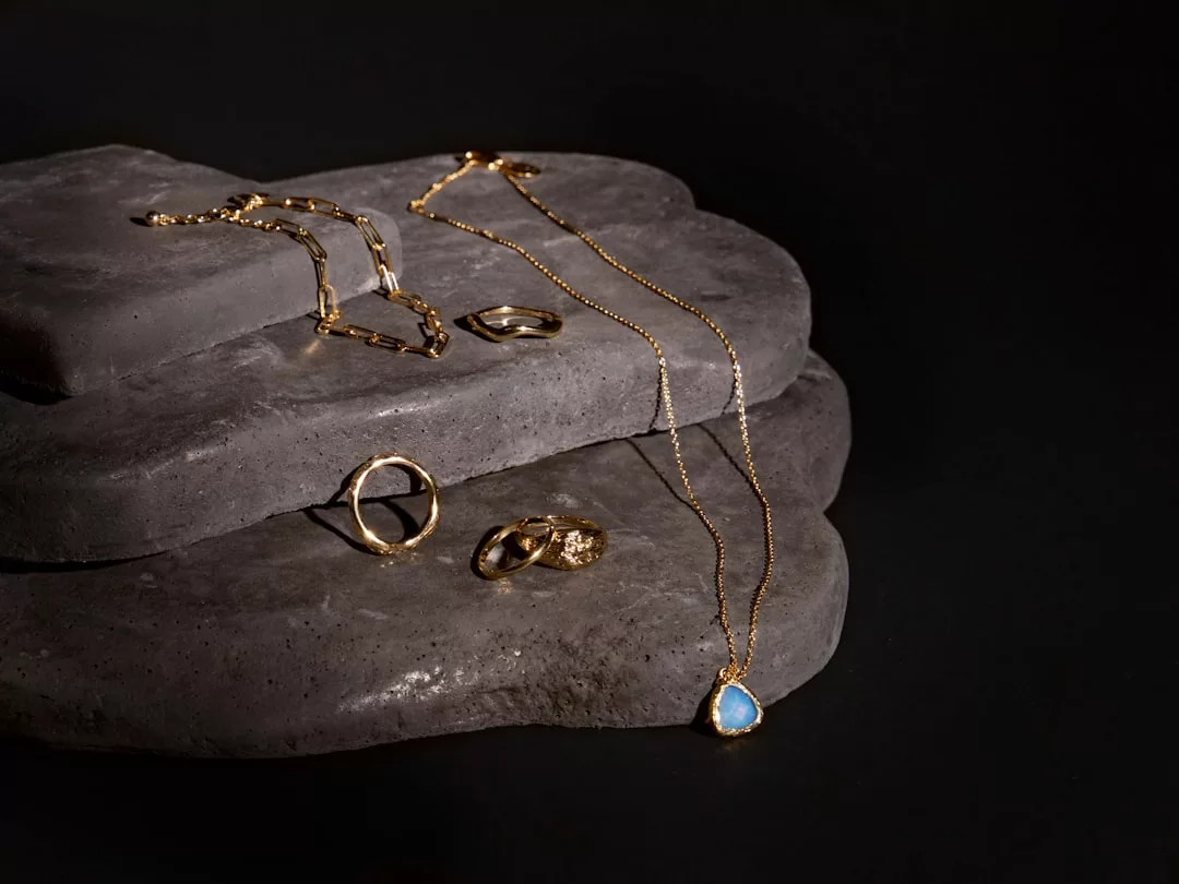 Der Goldschmuck von GLAMESiA, darunter Ringe, ein Armband und eine Halskette mit einem Anhänger aus blauem Stein, wird elegant auf geschichteten dunklen Steinplatten vor einem schwarzen Hintergrund präsentiert.