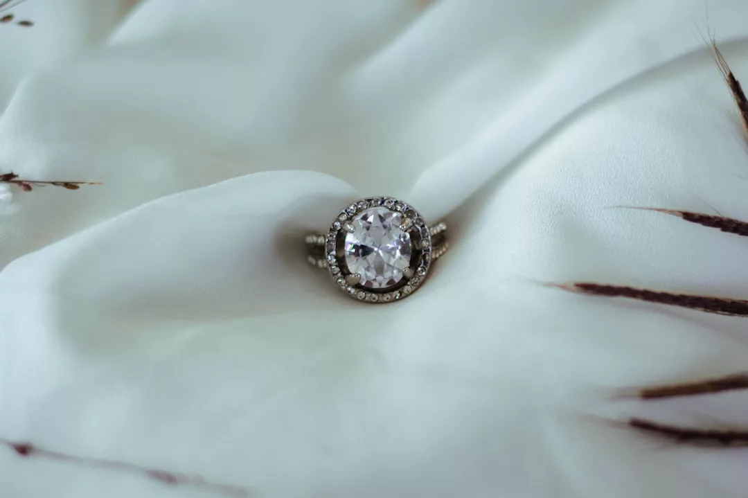 Ein GLAMESiA-Diamantring mit einem runden Mittelstein und einem Kranz kleinerer Diamanten ruht auf weißem Stoff mit zarten braunen Zweigen in der Nähe.