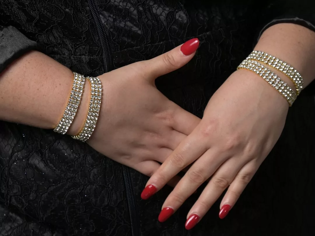 Eine Person mit rot lackierten Nägeln trägt an beiden Handgelenken goldene und diamantene Armreifen von GLAMESiA, die vor einem Hintergrund aus schwarzem, strukturiertem Stoff gezeigt werden.