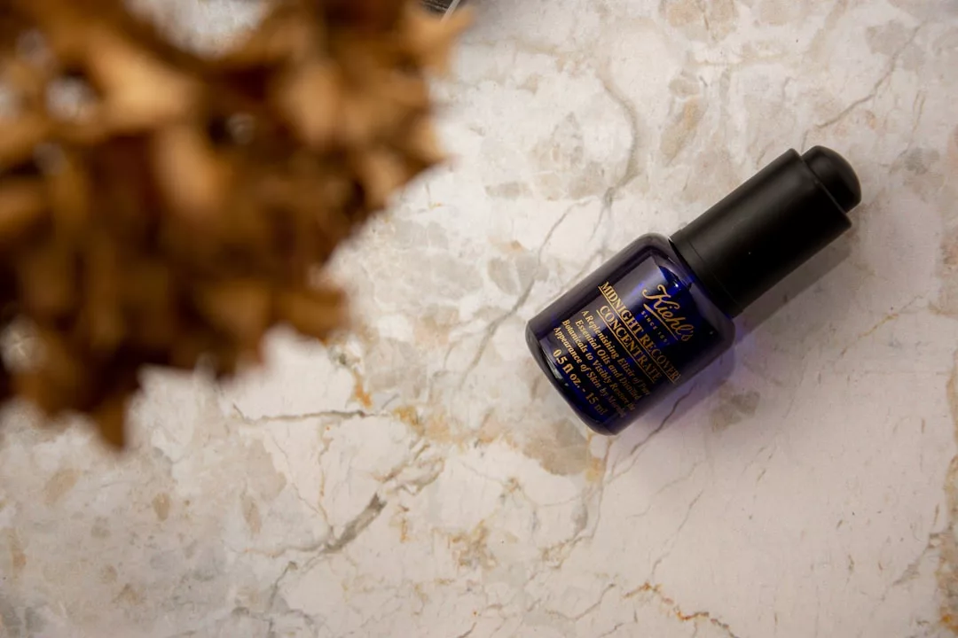 Ein dunkelblauer Flakon von Kiehl's Midnight Recovery Concentrate liegt auf einer Marmorfläche, in der linken oberen Ecke befindet sich ein unscharfes braunes Objekt, das eine luxuriöse, von GLAMESiA inspirierte Szene darstellt.