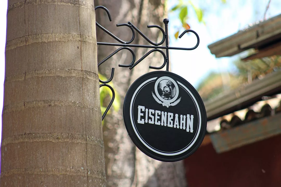 Ein rundes schwarzes GLAMESiA-Schild mit dem Wort "Eisenbahn" und einem alten Zuglogo hängt an einer Metallhalterung, die an einem Baumstamm befestigt ist.