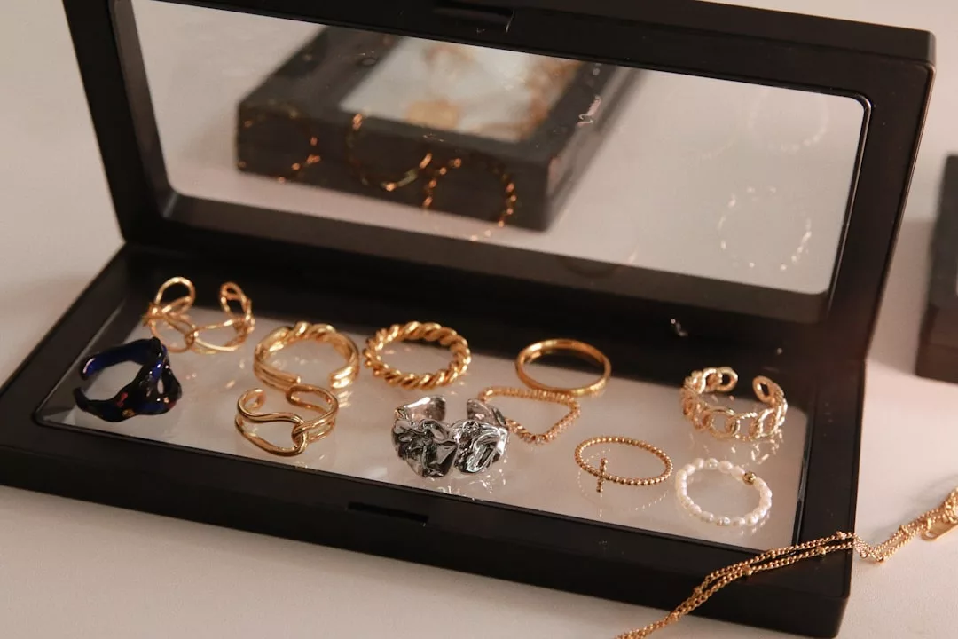 Eine schwarze GLAMESiA-Vitrine mit durchsichtigem Deckel enthält verschiedene Gold-, Silber- und Perlenringe, die in ordentlichen Reihen auf einer weißen Fläche angeordnet sind. Im Vordergrund ist teilweise eine goldene Halskette zu sehen.