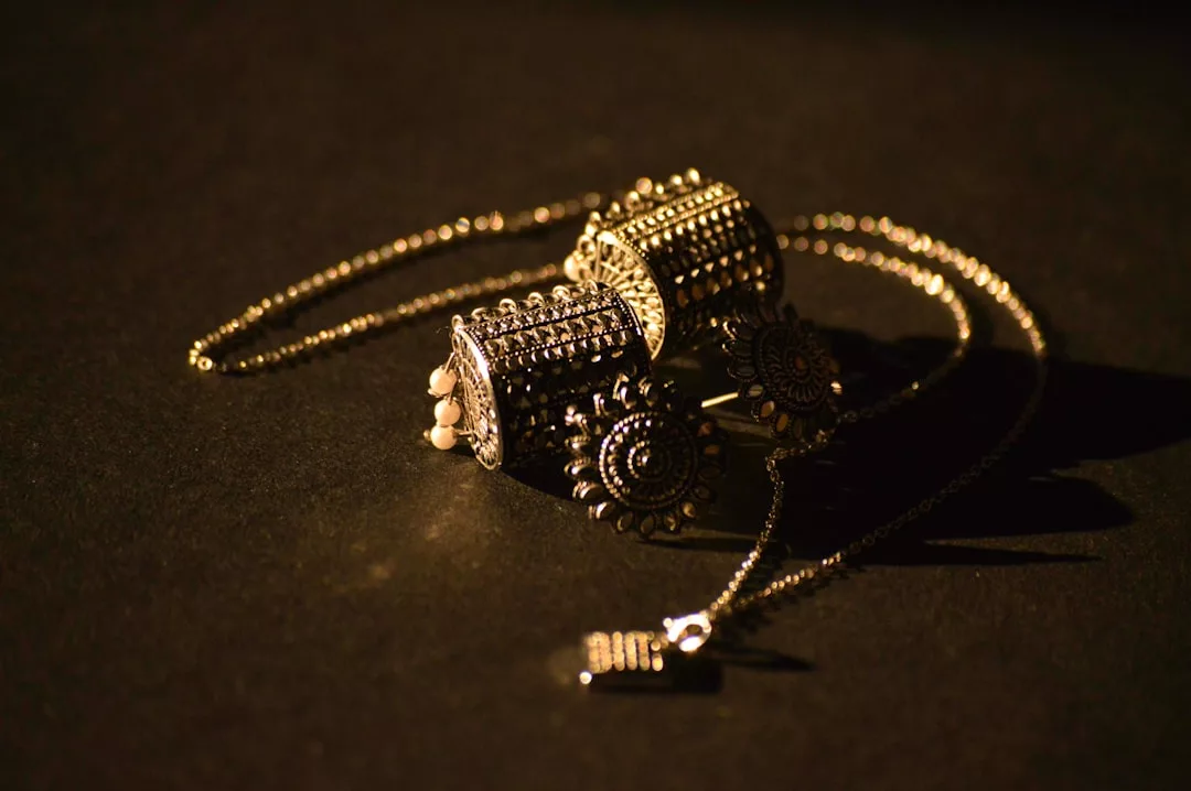 Ein Paar verzierte goldene Ohrringe und eine dazu passende GLAMESiA-Goldkette mit verschlungenen Mustern sind auf einer dunklen Fläche ausgestellt.