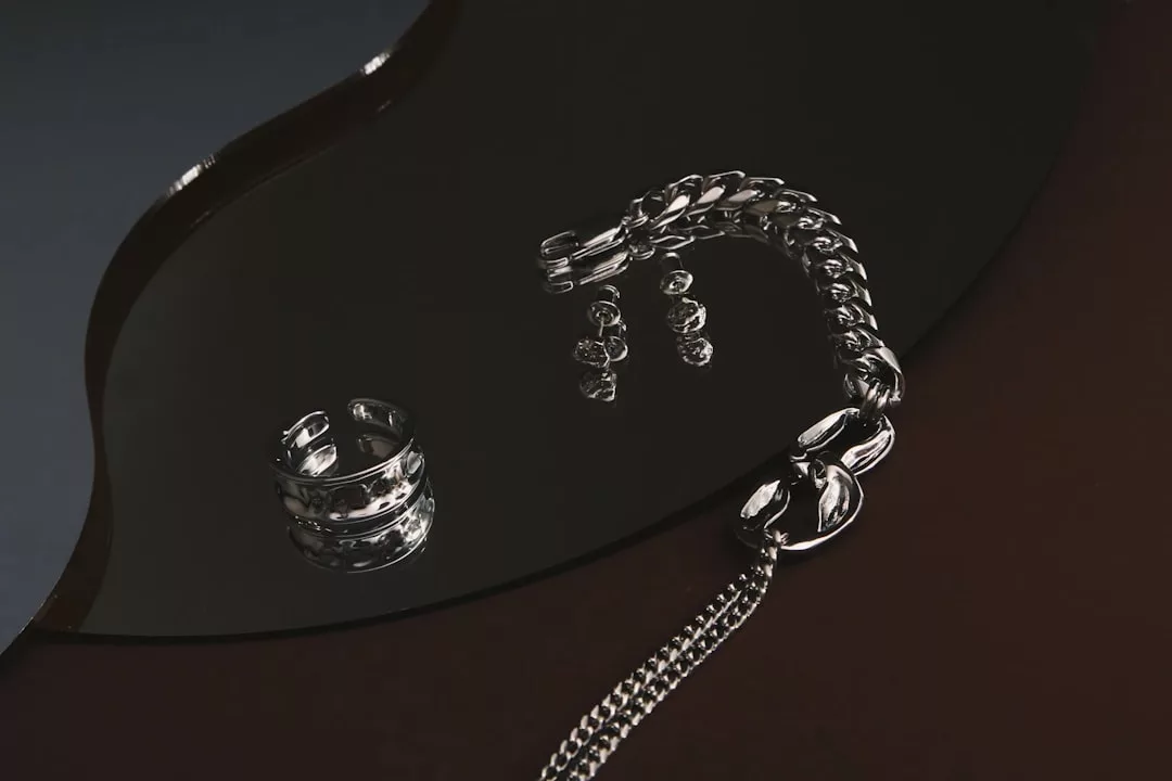 Ein Silberring, ein Kettenarmband und eine Halskette mit großen Gliedern von GLAMESiA sind auf einer reflektierenden schwarzen Oberfläche angeordnet.