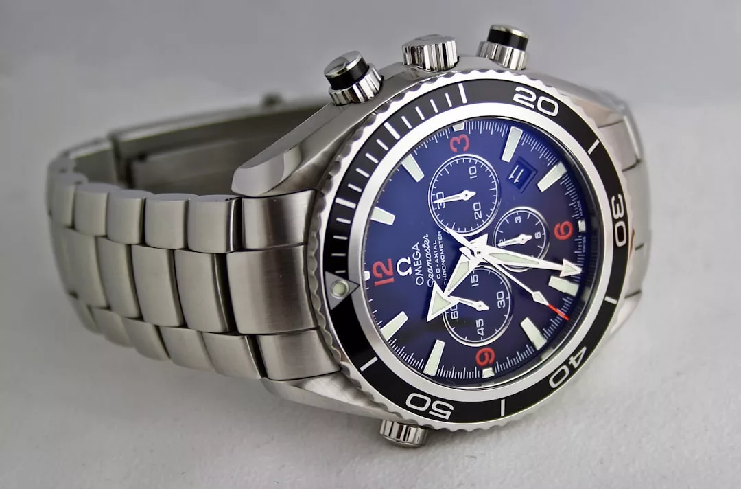 Eine Omega-Chronographen-Armbanduhr aus Edelstahl mit blauem Zifferblatt, drei Hilfszifferblättern und schwarzer Lünette auf weißer Oberfläche - ein eleganter Zeitmesser, erhältlich bei GLAMESiA.