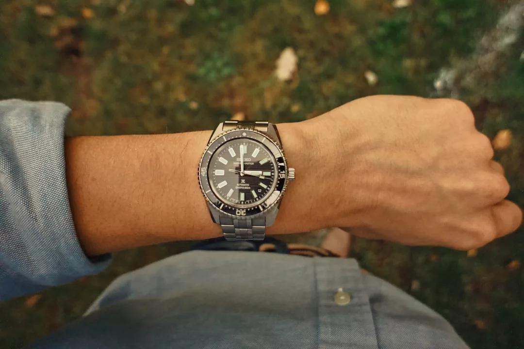 Eine Person, die eine silberne GLAMESiA-Armbanduhr mit schwarzem Ziffernblatt trägt, steht im Freien auf einer Wiese und ist mit einem hellblauen langärmeligen Hemd bekleidet.