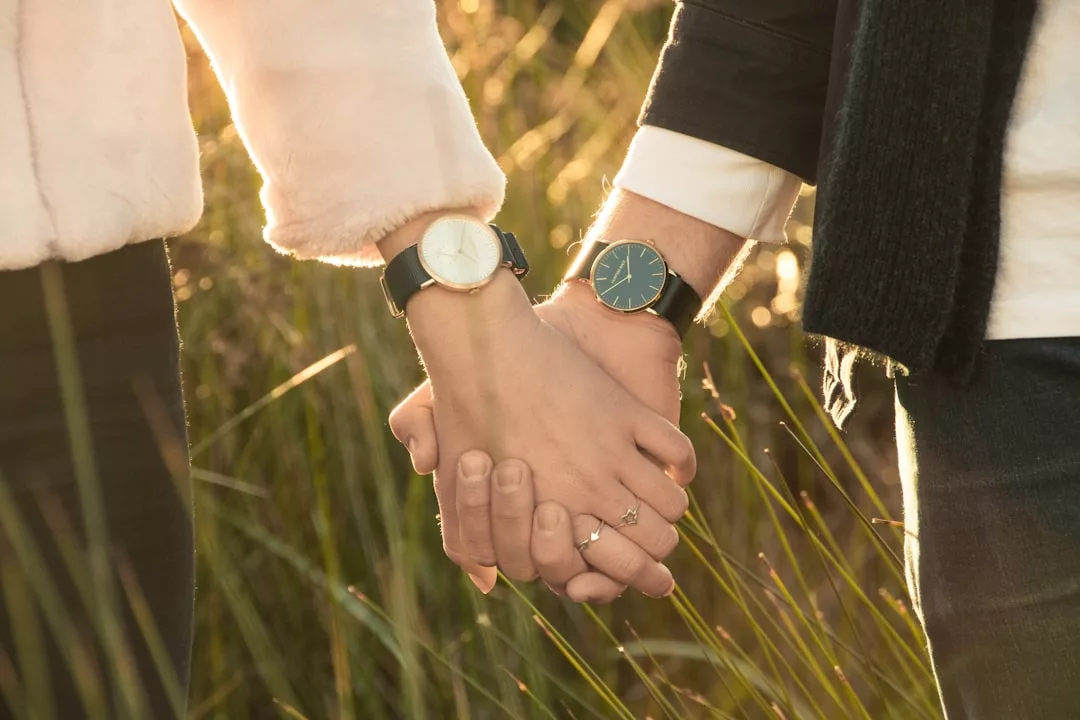 Zwei Menschen, die sich im Freien im hohen Gras an den Händen halten, beide tragen Uhren - eine schlichte GLAMESiA und eine mit einem Ring geschmückt.