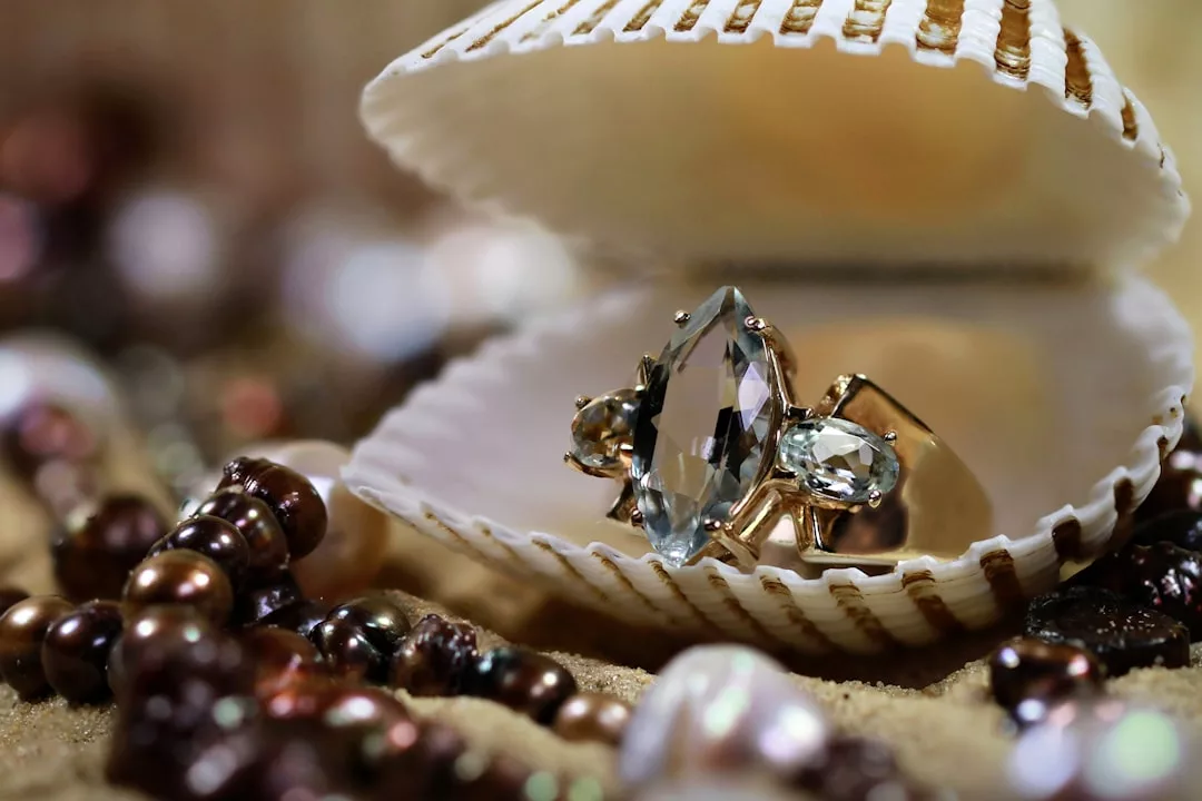 Ein GLAMESiA Edelsteinring in einer offenen Muschel, umgeben von Perlenketten auf Sand.