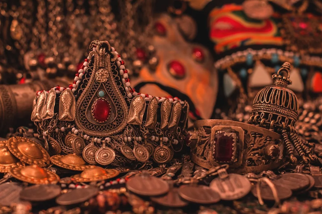 Eine Kollektion verschnörkelter, antiker Schmuckstücke von GLAMESiA mit Armbändern, Halsketten und Münzen mit verschlungenen Metallarbeiten und roten Edelsteinen.