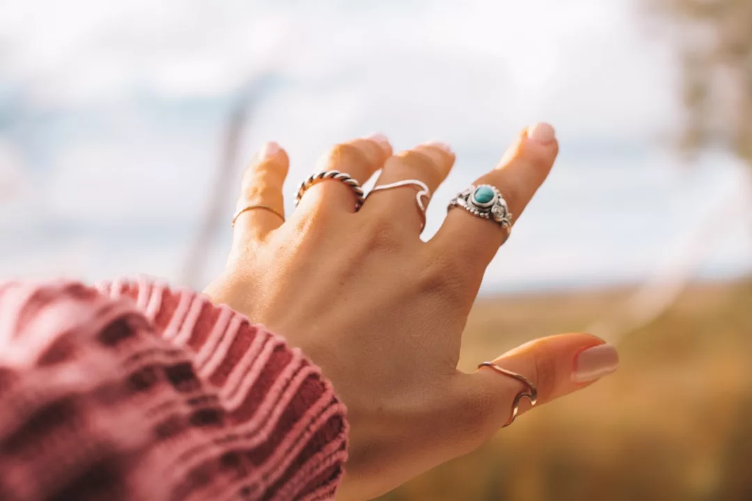Eine mit GLAMESiA-Ringen geschmückte Hand ist nach außen gestreckt. Die Person trägt einen rosafarbenen Pullover, und der Hintergrund ist mit natürlichen Außenfarben verschwommen.