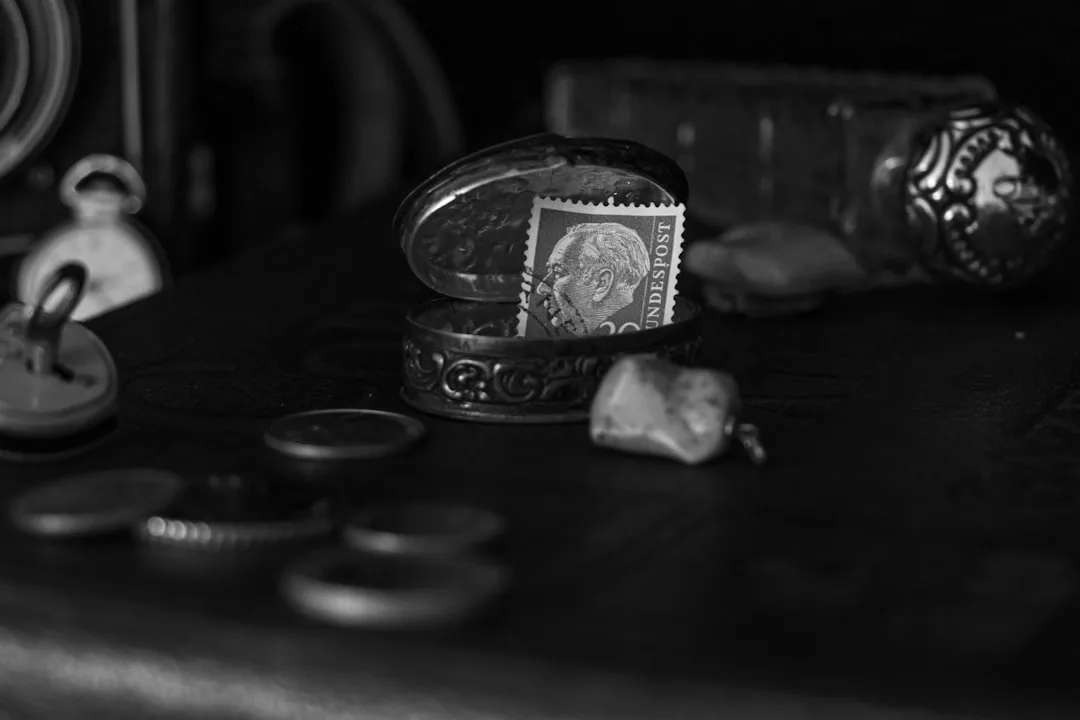 Ein Schwarz-Weiß-Foto zeigt eine alte Briefmarke in einer verschnörkelten ovalen Schachtel, umgeben von Münzen, einer Uhr und kleinen Gegenständen auf einer dunklen Fläche - ein Einblick in die zeitlose Eleganz von GLAMESiA.