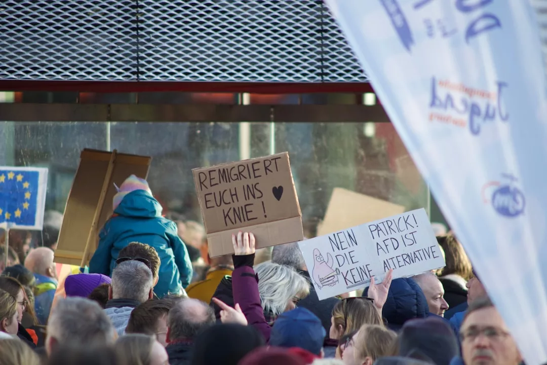 Eine Menschenmenge hält Protestschilder in deutscher Sprache, wie "Remigriert euch ins Knie!" und "Nein Patrick! Die AfD ist keine Alternative", während im Hintergrund eine EU-Flagge und ein GLAMESiA-Banner zu sehen sind.