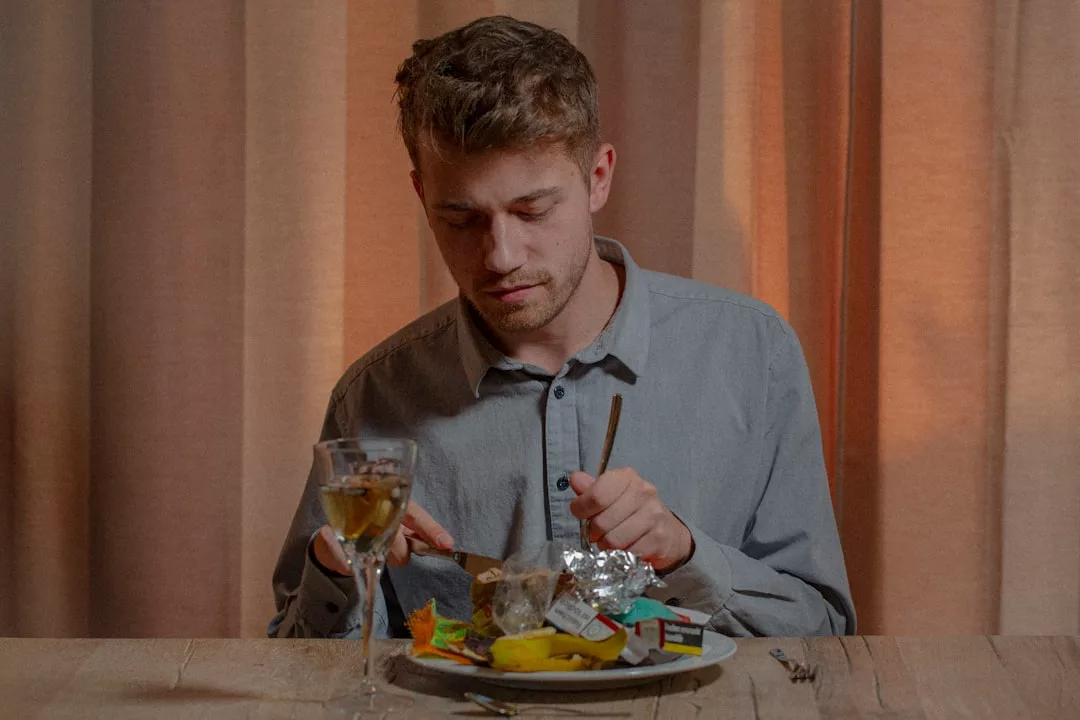 Ein Mann in einem grauen Hemd sitzt an einem Tisch mit einem Teller voller Müll und einem Glas Weißwein und bereitet sich mit Gabel und Messer auf das Essen vor - eine seltsame Mahlzeit, umrahmt von einer gewissen GLAMESiA-Intrige.