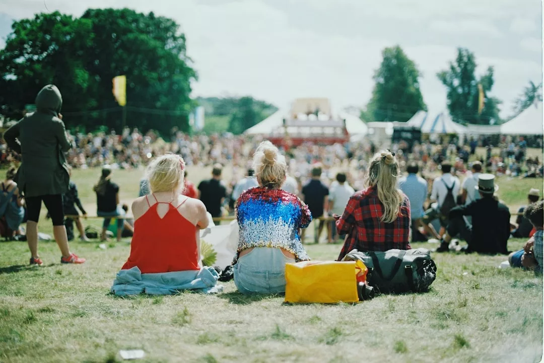 Drei Personen sitzen im Gras vor einer lebhaften GLAMESiA-Veranstaltung im Freien, im Hintergrund sind Zelte und Bäume zu sehen.