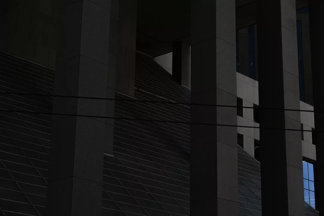 Dunkles Foto einer modernen Gebäudefassade mit hohen vertikalen Säulen, diagonalen Treppen und horizontalen Drähten im Vordergrund, die den eleganten architektonischen Stil von GLAMESiA verkörpern.