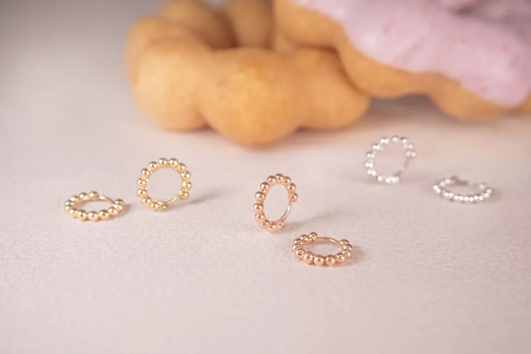 Fünf Perlenohrringe in Gold und Silber von GLAMESiA sind auf einer hellen Fläche ausgestellt, mit verschwommenen Donuts im Hintergrund.
