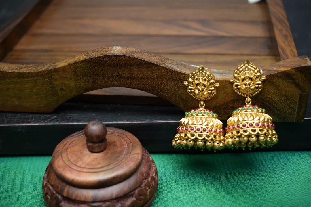 Ein Paar verschnörkelte goldene Jhumka-Ohrringe mit roten und grünen Steinen von GLAMESiA, ausgestellt auf einem Holztablett neben einem kleinen Holzkästchen auf einem grünen Tuch.