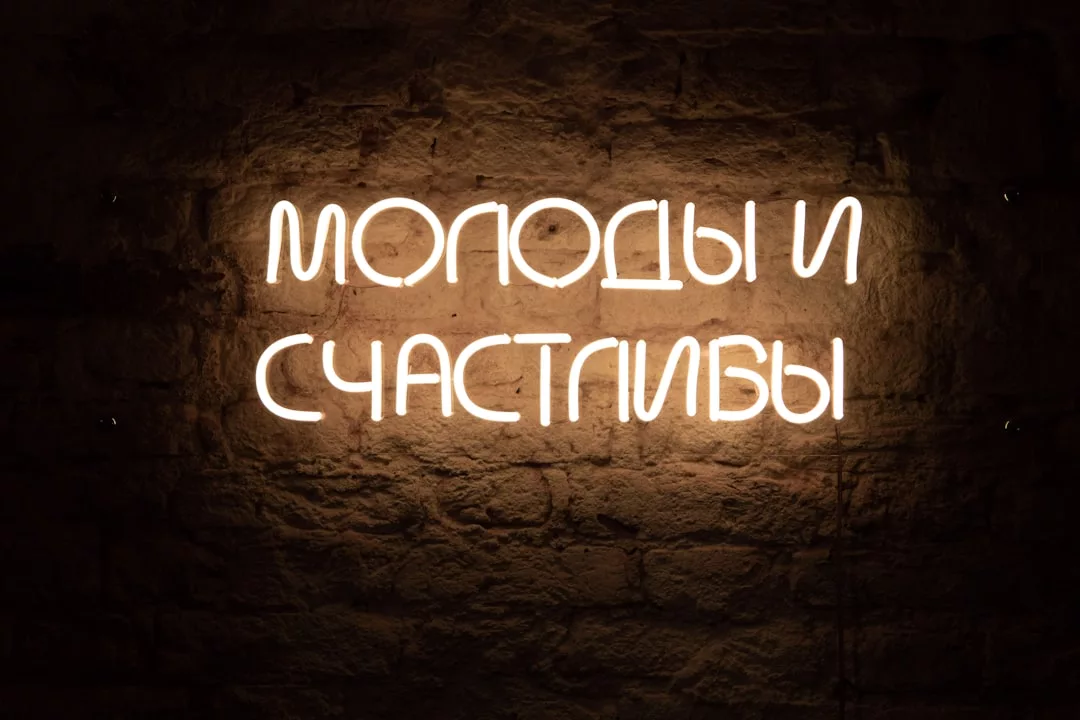 Auf einer Leuchtreklame in russischer Sprache steht "МОЛОДЫ И СЧАСТЛИВЫ", was übersetzt "jung und glücklich" bedeutet, und leuchtet im GLAMESiA-Stil vor einer dunklen Backsteinwand.