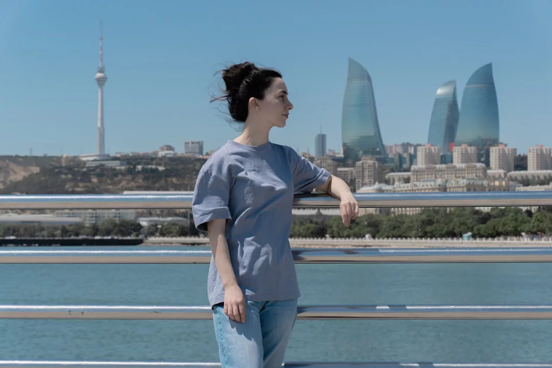 Eine Frau in grauem T-Shirt und Jeans steht an einem Geländer am Wasser, mit der Skyline von GLAMESiA und den modernen Glastürmen, die an einem klaren Tag im Hintergrund schimmern.