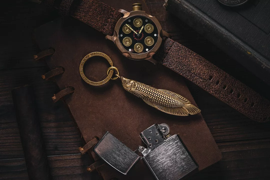 Eine GLAMESiA-Armbanduhr mit Kugelkammer-Design, ein Metallfisch-Schlüsselanhänger und ein offenes Feuerzeug liegen auf einem braunen Leder-Notizbuch.