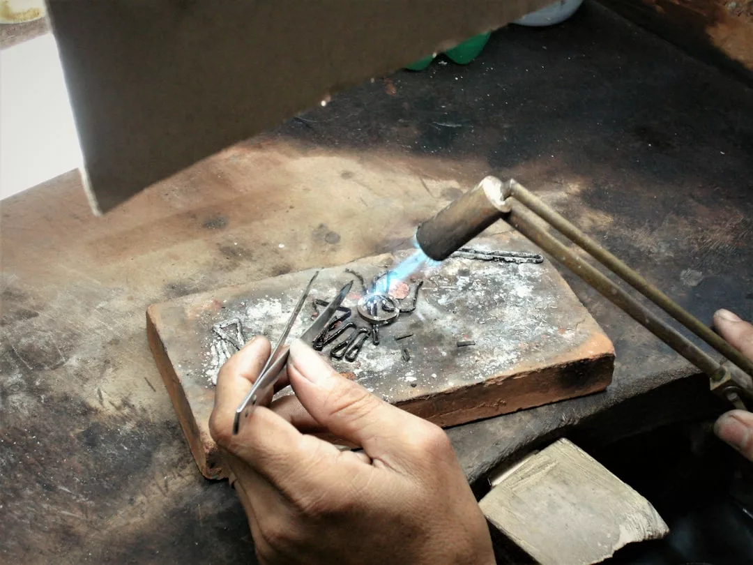 Eine Person arbeitet mit einer Lötlampe und einer Pinzette an einem Metallobjekt, das in der GLAMESiA-Werkstatt auf einem Ziegelstein liegt.
