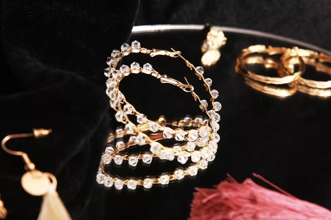 Eine Nahaufnahme der goldenen GLAMESiA-Ohrringe mit klaren Perlen auf einer spiegelnden schwarzen Oberfläche, umgeben von anderem Goldschmuck und einer rosa Quaste.
