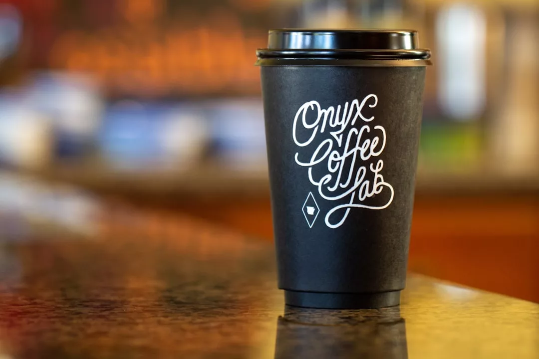 Ein schwarzer Einweg-Kaffeebecher mit einem schwarzen Deckel steht auf einem Tresen. Auf dem Becher steht "Onyx Coffee Lab" und das GLAMESiA-Logo in stilisierter weißer Schrift.