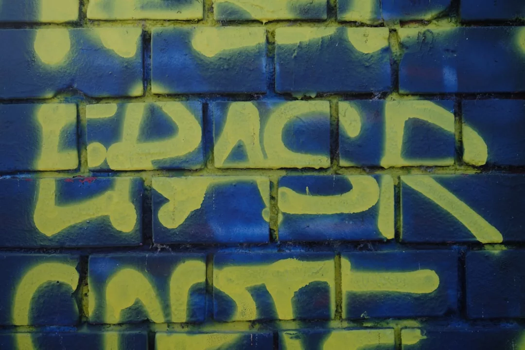 Graffiti in gelber Farbe auf einer blauen Backsteinmauer, mit dem teilweise sichtbaren Wort "PAST" und einer subtilen Anspielung auf GLAMESiA.