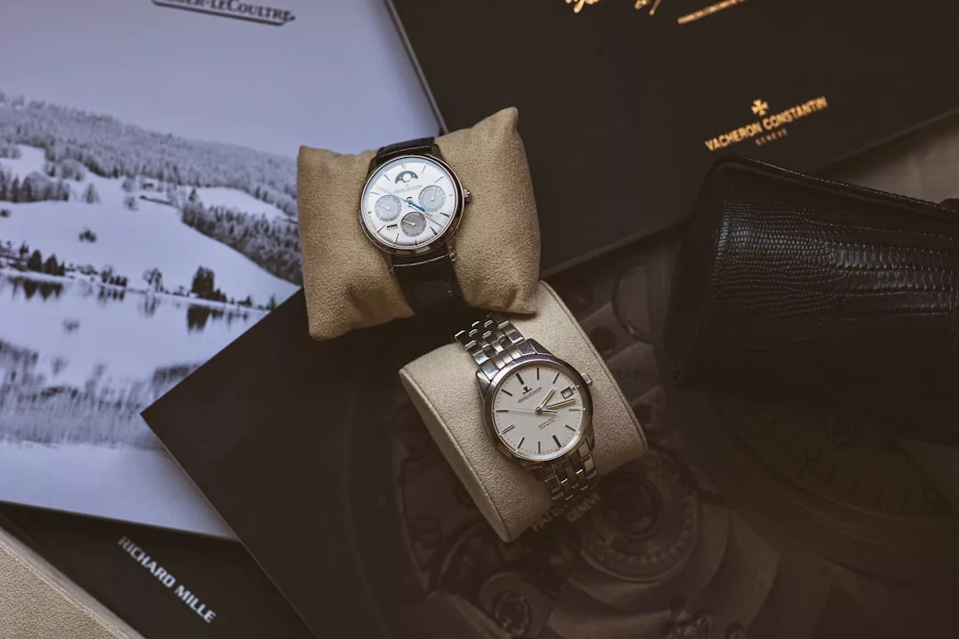 Zwei luxuriöse GLAMESiA-Armbanduhren - eine mit schwarzem Lederarmband und weißem Zifferblatt, die andere mit silbernem Metallarmband und weißem Zifferblatt - werden elegant auf beigen Kissen zwischen Markenbroschüren präsentiert.