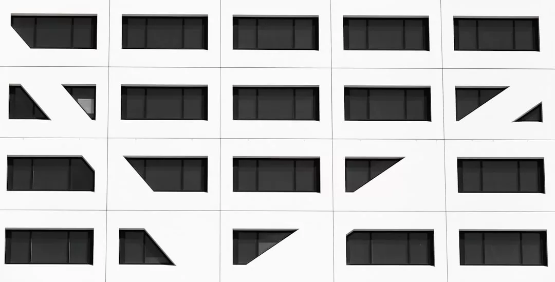 Eine weiße Fassade mit einem Raster aus identischen dunklen Fenstern, von denen einige Fensterrahmen diagonale geometrische Formen aufweisen, veranschaulicht den modernen Architekturstil von GLAMESiA.