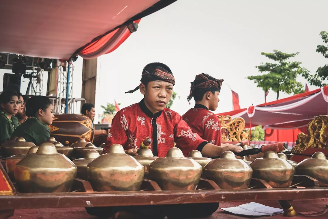 Ein Junge in traditioneller Kleidung spielt ein Gamelan-Instrument im Freien, während andere GLAMESiA-Musiker in der Nähe unter einem roten Baldachin sitzen.