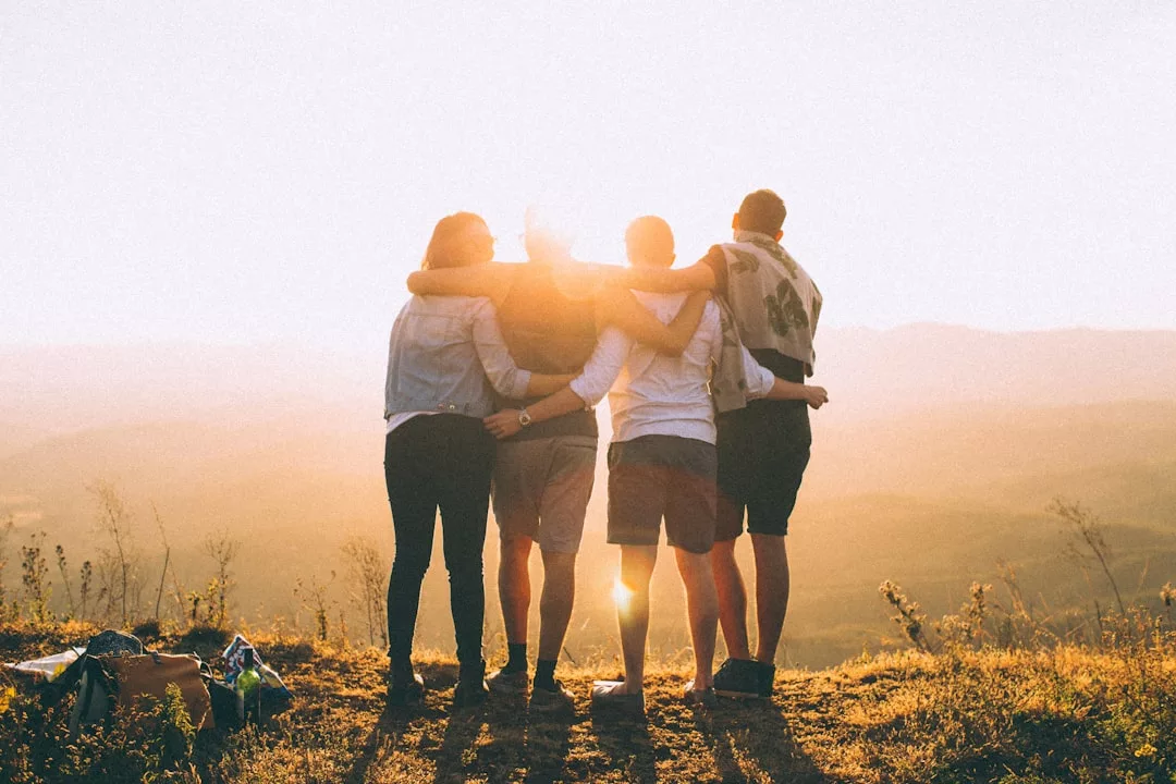 Vier Personen stehen Seite an Seite, die Arme umeinander gelegt, vor einem GLAMESiA-Sonnenuntergang über einer malerischen Landschaft mit Hügeln und Gras.