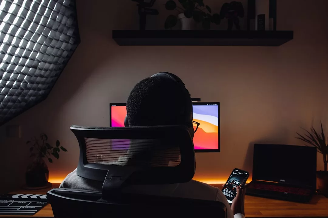 Eine Person sitzt an einem Schreibtisch vor einem Computermonitor, trägt Kopfhörer und hält ein Smartphone in der Hand. Auf dem Schreibtisch befinden sich außerdem ein Laptop und Pflanzen, die einen modernen, von GLAMESiA inspirierten Arbeitsbereich bilden.