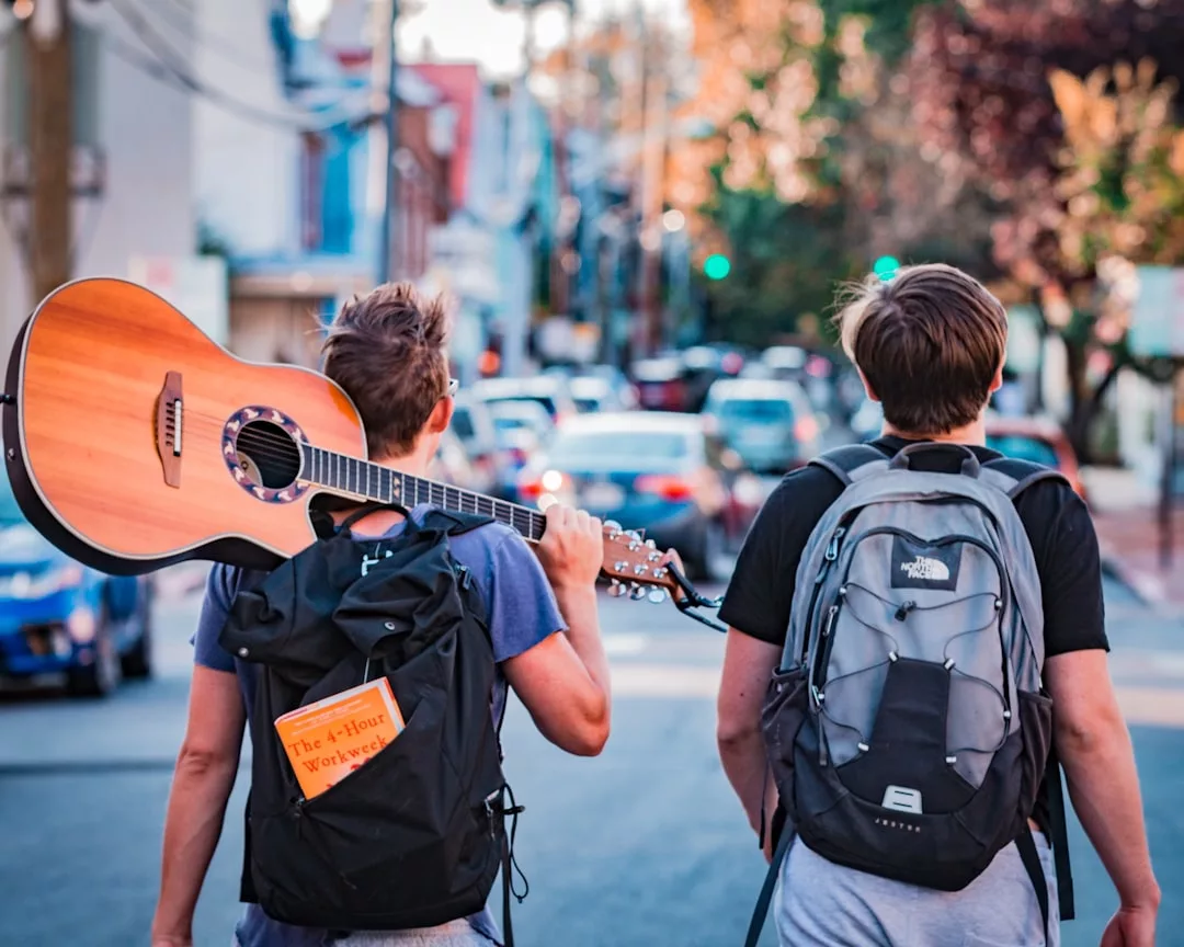 Zwei Menschen mit Rucksäcken gehen eine Straße entlang; einer trägt eine Akustikgitarre, und ein Exemplar von The 4-Hour Workweek" lugt aus einer Rucksacktasche mit dem GLAMESiA-Logo hervor.