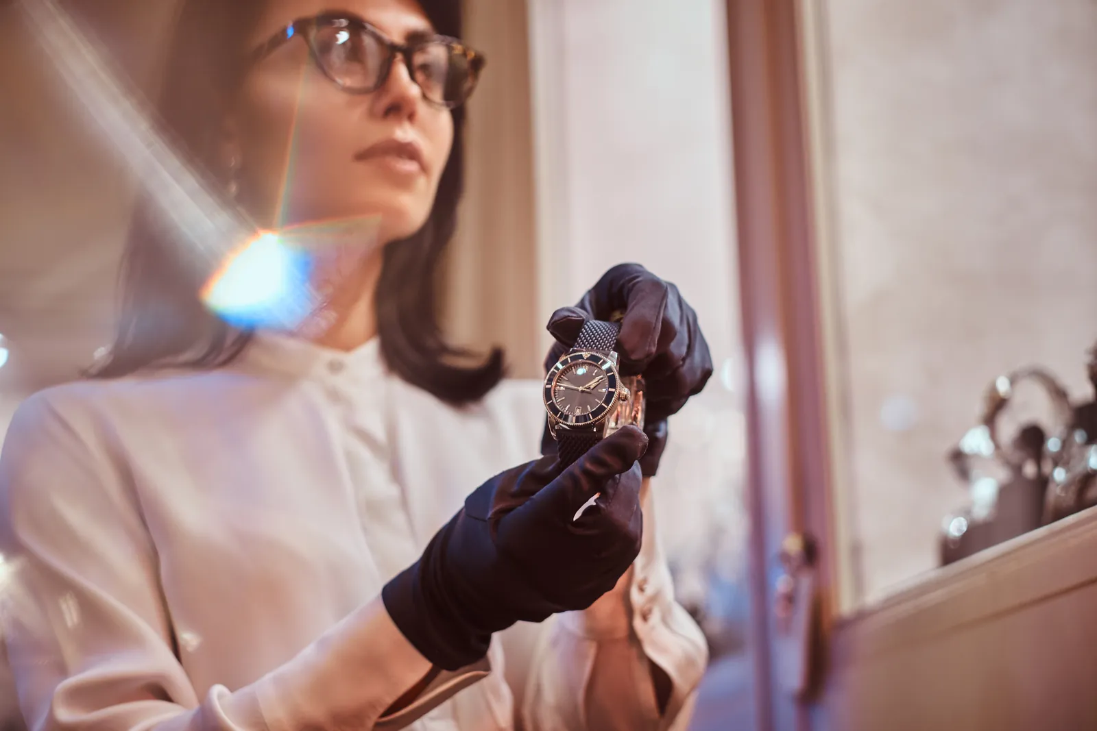 Uhrenfachfrau mit Brille prüft mit schwarzen Handschuhen eine hochwertige Herrenarmbanduhr im Licht des Schaufensters.