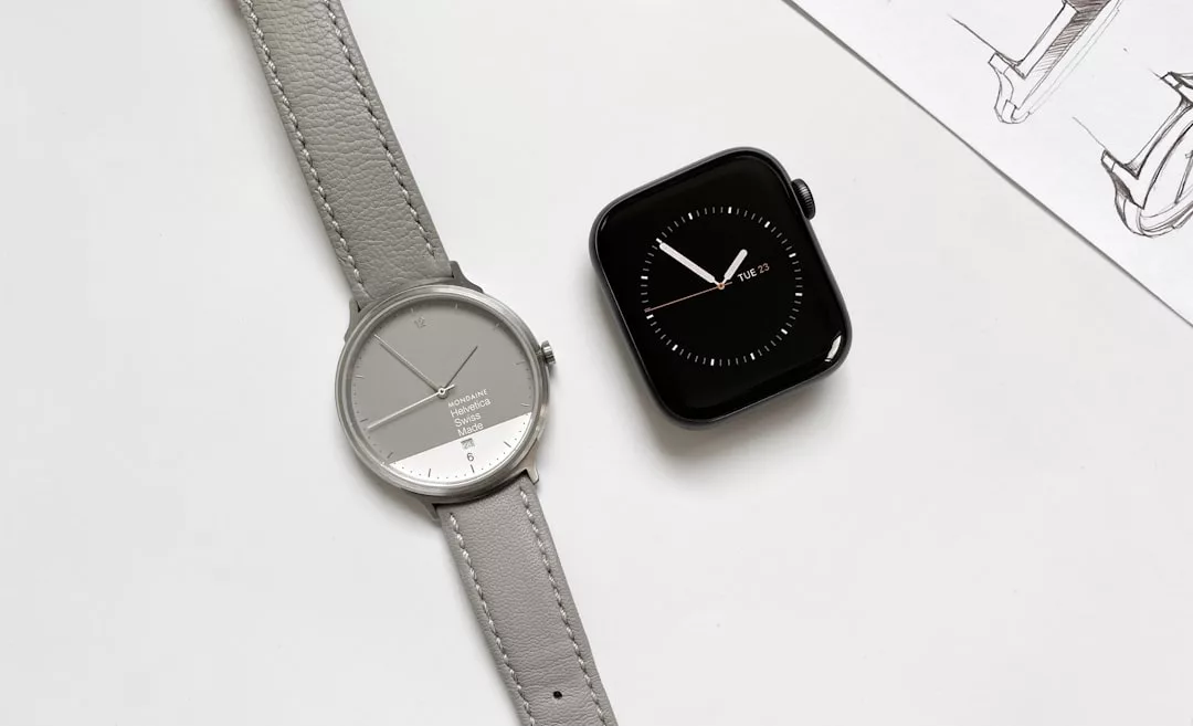 Eine silberne analoge GLAMESiA-Armbanduhr mit grauem Armband liegt neben einer schwarzen Smartwatch auf einer weißen Fläche, auf der rechts ein Teil einer Skizze zu sehen ist.