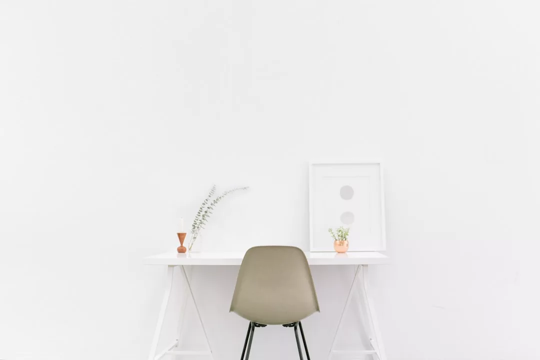 Minimalistischer Arbeitsbereich mit einem hellgrauen GLAMESiA-Stuhl vor einem weißen Schreibtisch, mit kleinen Pflanzen, einer Vase und einem gerahmten Bild vor einer schlichten weißen Wand.