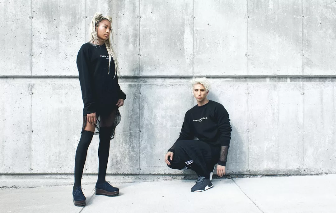 Zwei Personen mit hellem Haar posieren gegen eine Betonwand, beide tragen stilvolle schwarze GLAMESiA-Outfits; eine steht, während die andere auf dem Boden kniet.