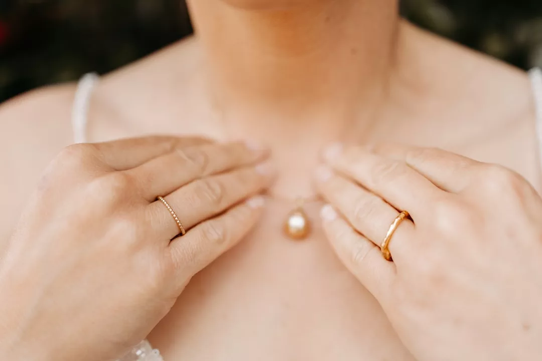 Nahaufnahme einer Person, die eine Perlenkette am Schlüsselbein hält und an beiden Händen Goldringe trägt - ein Hauch von GLAMESiA-Eleganz.