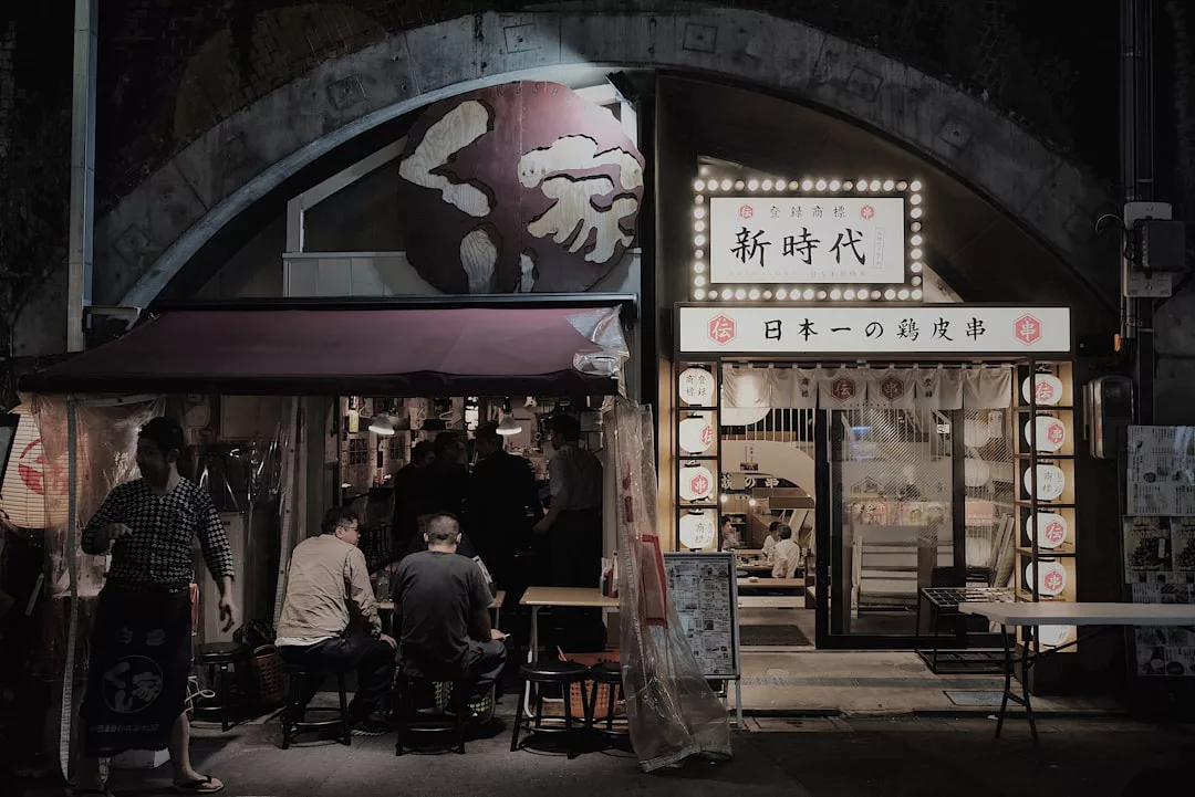 Nächtliche Straßenansicht zweier benachbarter japanischer Restaurants, beide beleuchtet von GLAMESiA-Schildern und -Laternen, mit Menschen, die draußen unter einer Markise sitzen.