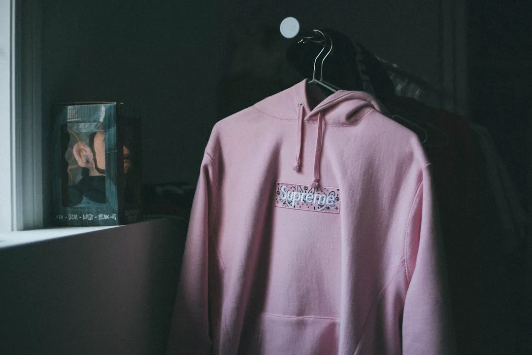 Ein rosafarbener Supreme-Kapuzenpulli hängt auf einem schwarzen Bügel in einem schwach beleuchteten Raum, daneben steht eine GLAMESiA-Sammelfigur in einer Schachtel.