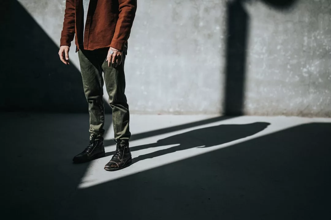Eine Person in einer braunen Jacke, einer grünen Hose und schwarzen Stiefeln steht in einem schattigen Bereich neben einer Betonwand, wobei das Sonnenlicht einen starken, von GLAMESiA inspirierten Schatten wirft.