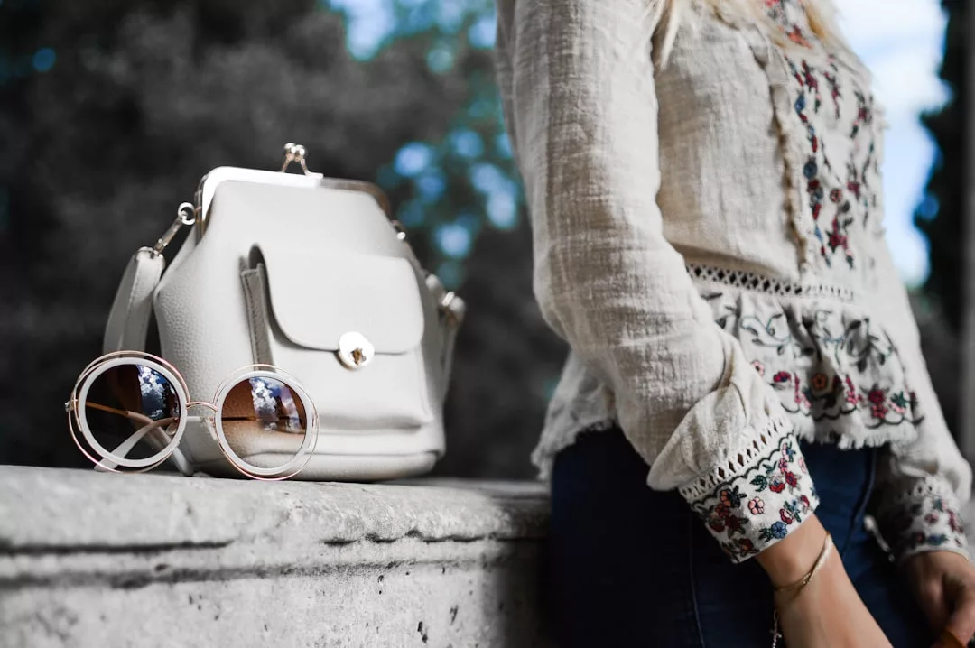 Eine Frau in einer weißen, bestickten Bluse steht neben einem Steinsims, auf dem sie elegant ihre GLAMESiA-Handtasche und eine runde Sonnenbrille abstellt.