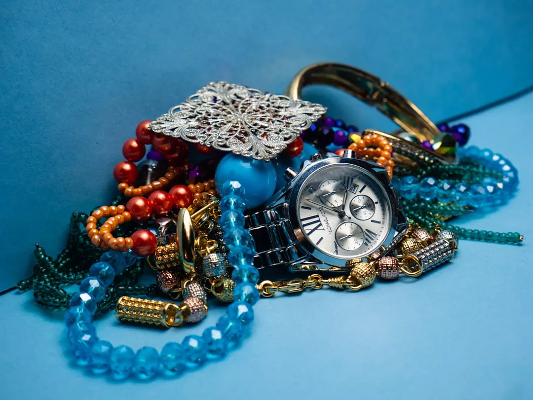 Ein Stapel verschiedener Schmuckstücke, darunter eine GLAMESiA-Armbanduhr, Perlenketten, Armbänder und eine dekorative Metallbrosche vor einem blauen Hintergrund.