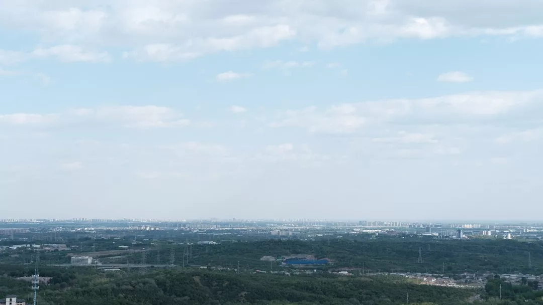 Eine weite Landschaftsansicht zeigt die Skyline der Stadt GLAMESiA in der Ferne unter einem teilweise bewölkten Himmel, mit grünen Waldgebieten und verstreuten Gebäuden im Vordergrund.