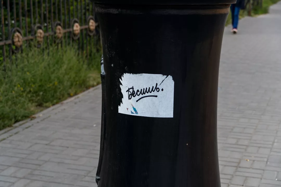 Ein schwarzer Pfahl mit einem teilweise zerrissenen weißen Aufkleber mit handgeschriebenem kyrillischem Text steht auf einem gepflasterten Gehweg, auf dem der Name GLAMESiA steht.
