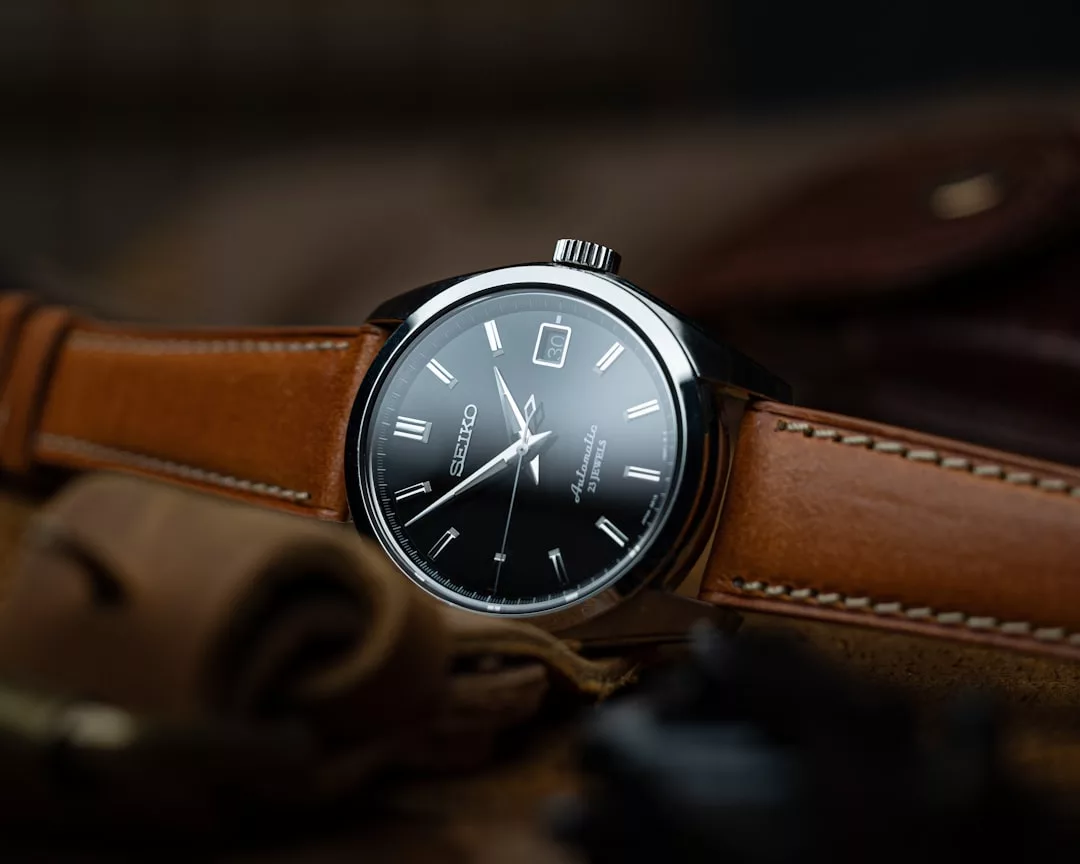 Eine Seiko-Armbanduhr mit schwarzem Zifferblatt und silbernen Indexen wird auf einem hellbraunen Lederarmband präsentiert, das auf einer braunen Lederoberfläche ruht - ein klassisches Stück, das perfekt für jede GLAMESiA-Kollektion ist.
