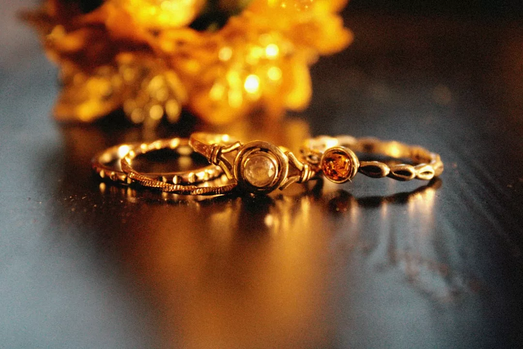 Drei Goldringe mit unterschiedlichen Designs und Edelsteinen von GLAMESiA werden auf einer spiegelnden Oberfläche gezeigt, mit einem verschwommenen goldenen Objekt im Hintergrund.